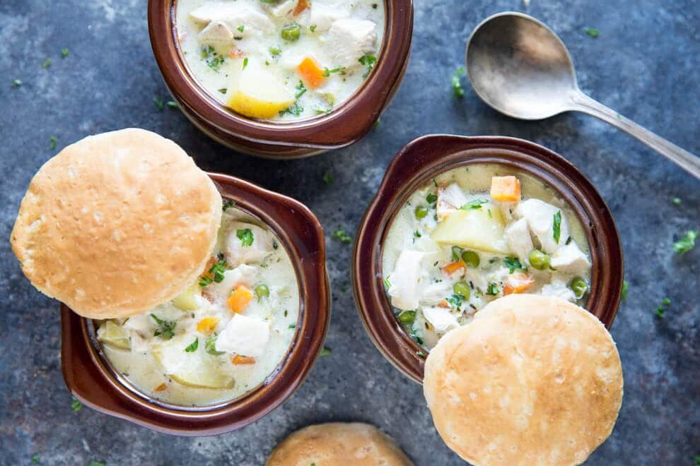 A portrait shaped photo of three bowls of chicken pot pie soup with carrots, peas, and potatoes in a creamy sauce with three freshly made biscuits vidisble. There is a spoon in the lower right hand corner. the bowls are brown on a blue background.