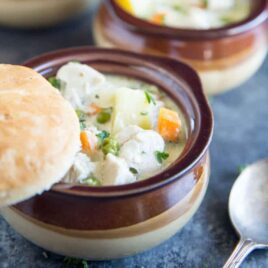 Bowl of creamy and delicious Chicken Pot Pie soup with carrot, peas, potatoes and chunks of white chicken meat in a creamy sauce with a freshly made biscut balancing on the top of the brown bowl. There is a spoon in the foreground, and a second bowl of soup in the background.
