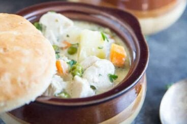 Bowl of creamy and delicious Chicken Pot Pie soup with carrot, peas, potatoes and chunks of white chicken meat in a creamy sauce with a freshly made biscut balancing on the top of the brown bowl. There is a spoon in the foreground, and a second bowl of soup in the background.