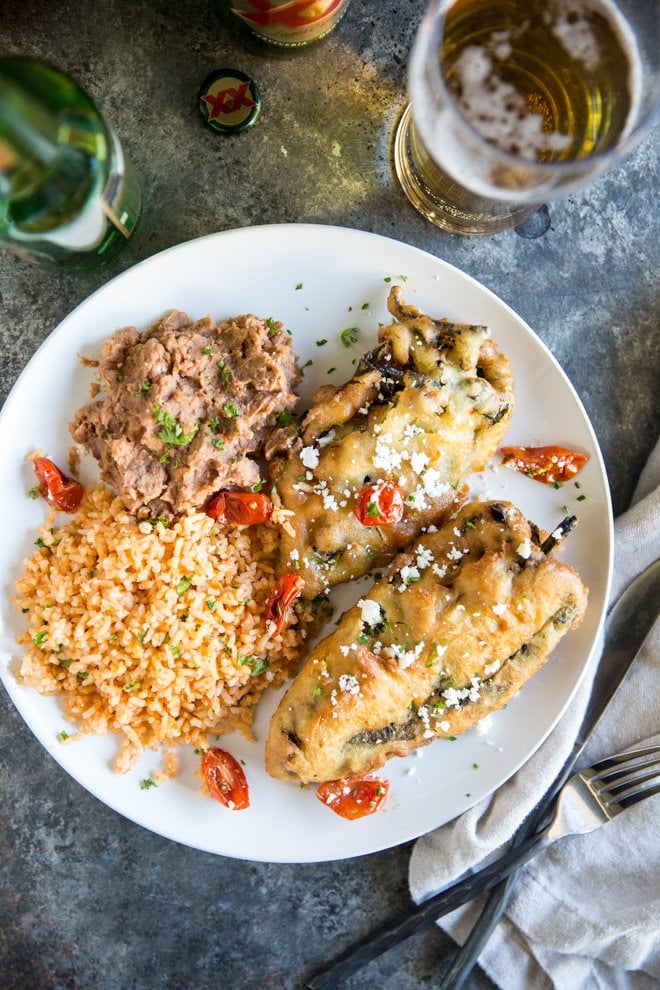 Overhead shot of two freshly fried chile rellenos with Mexican Rice and refried beans on a white plate with Dos Equis beer bottle in the background. Half the beer is poured into a glass.