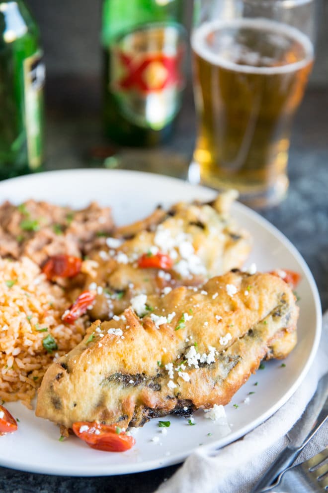 Two freshly fried chile rellenos with Mexican Rice and refried beans on a white plate with Dos Equis beer bottle in the background. Half the beer is poured into a glass.