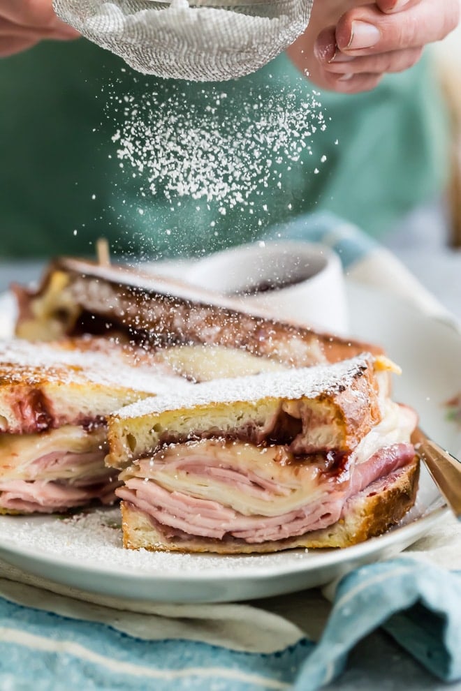 A monte cristo sandwich on a white platter being dusted with powdered sugar.