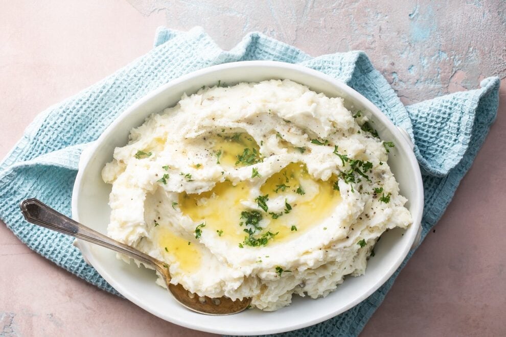 Make ahead mashed potatoes with melted butter in a white serving dish with a spoon on a blue towel.