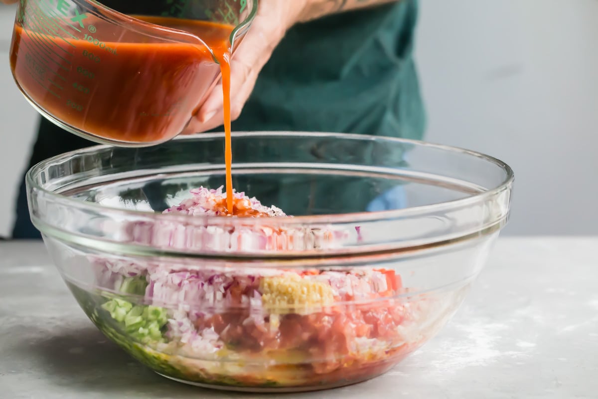 Someone pouring tomato juice into a bowl of vegetables.