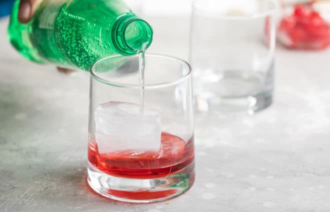 Grenadine and an ice cube in a clear glass with sprite being poured into it from a bottle.