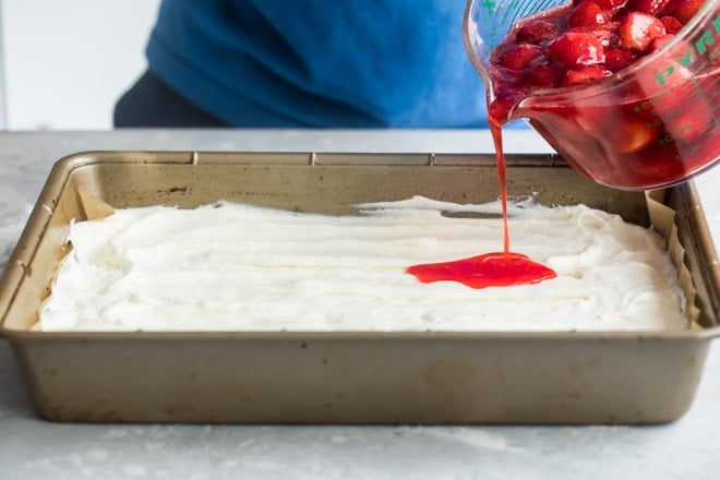 Strawberries being poured onto the white filling into a tan baking pan.