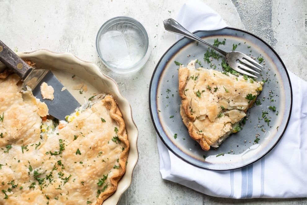 Turkey pot pie in a white pie pan and a piece of turkey pot pie on a blue plate with a fork and a clear glass of water.