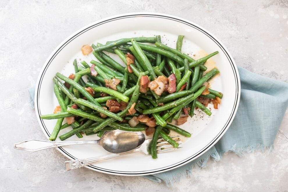 Green beans with bacon on a white platter with a serving spoon and fork.