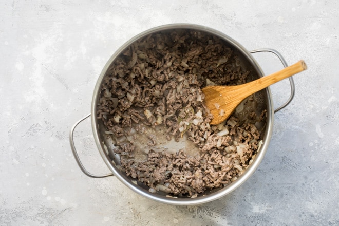 Ground beef and onions cooking in a silver pot with a wooden spoon.