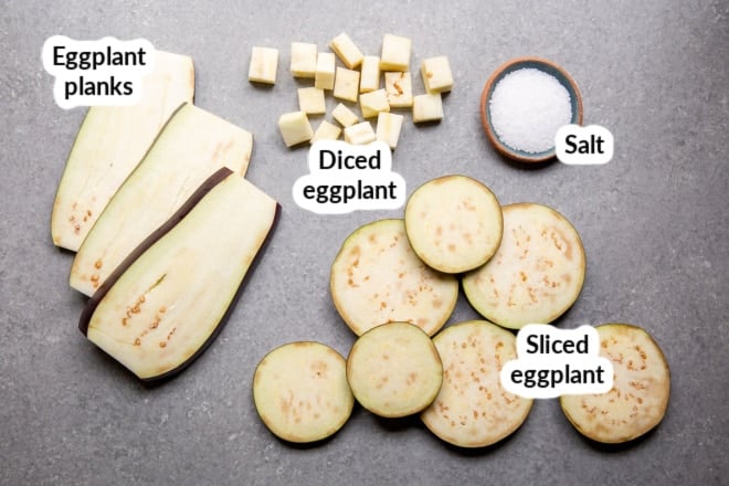 Eggplant cut into planks, slices, and cubes on a gray board.
