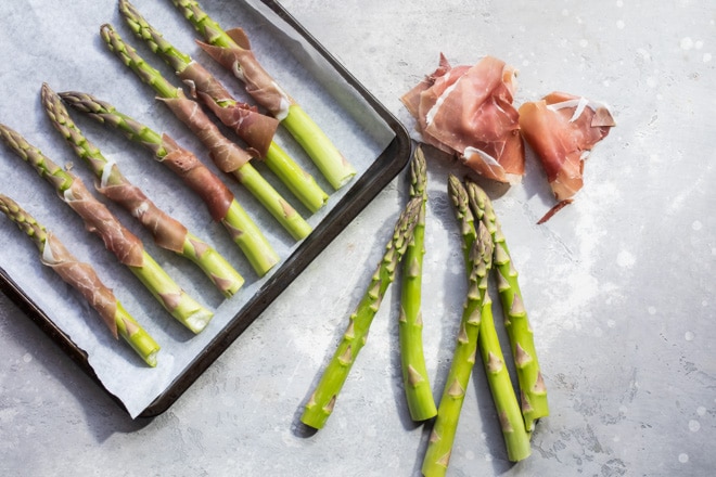 Prosciutto wrapped asparagus on a baking sheet with asparagus and prosciutto on the counter.