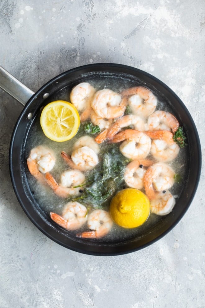Shrimp being poached in boiling water with lemon halves in a black skillet.