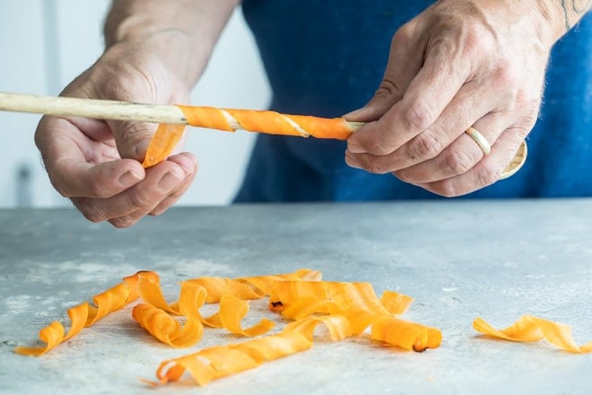 Someone curling long peels of carrot on a wooden stick.