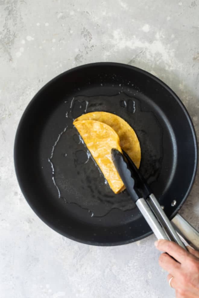 Someone flipping a corn tortilla in a black skillet with tongs.