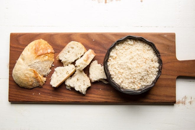 French bread, chunks of french bread, and a bowl of french breadcrumbs on a wood cutting board. 