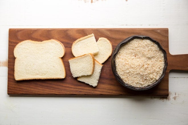 A slice of white bread, chunks of white bread, and a bowl of white breadcrumbs on a wood cutting board. 