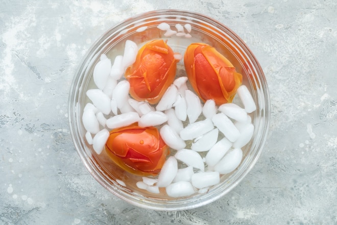 Three tomatoes being blanched in iced water in a clear bowl.