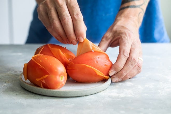 Someone peeling three tomatoes on a white plate.