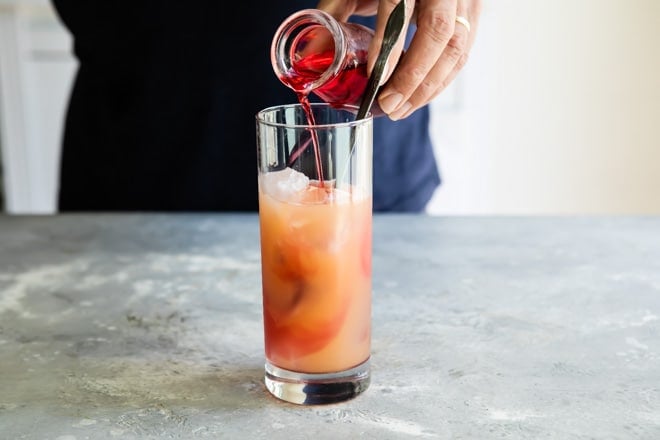 Someone adding grenadine into a seabreeze in a clear glass.