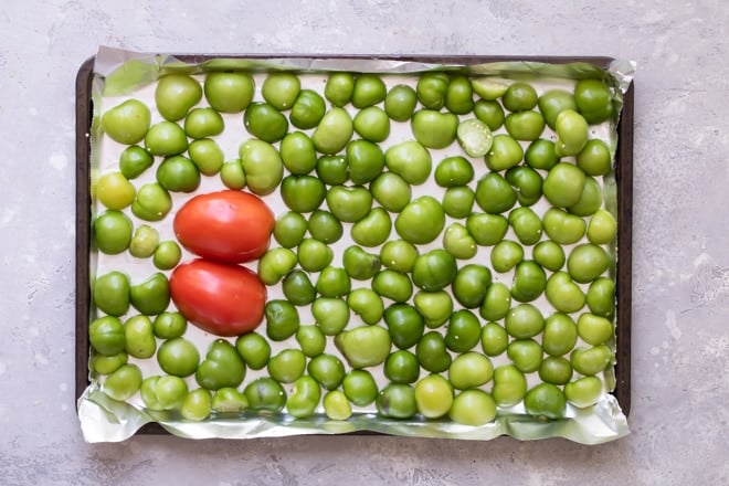 A halved tomato and halved green chilies on a baking sheet lined with tin foil.