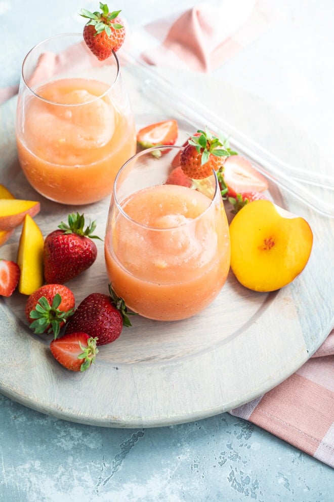 Two glasses of strawberry peach frose on a plate surrounded by slices of strawberries and peaches.