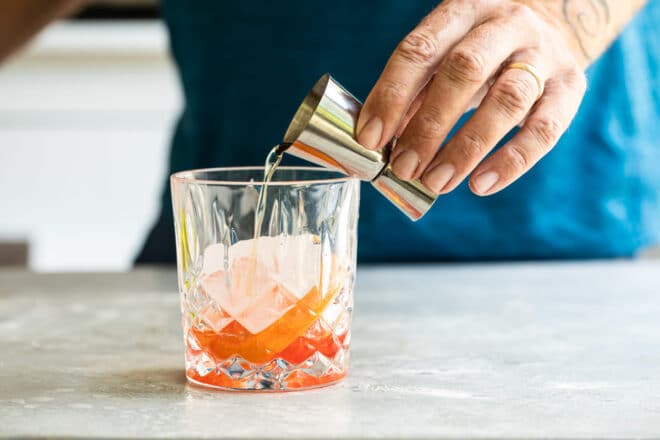 Brandy being poured into a clear glass with ice and red liquid in the bottom.