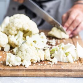 Raw cauliflower being chopped on a wooden cutting board.