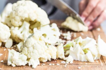 Raw cauliflower being chopped on a wooden cutting board.