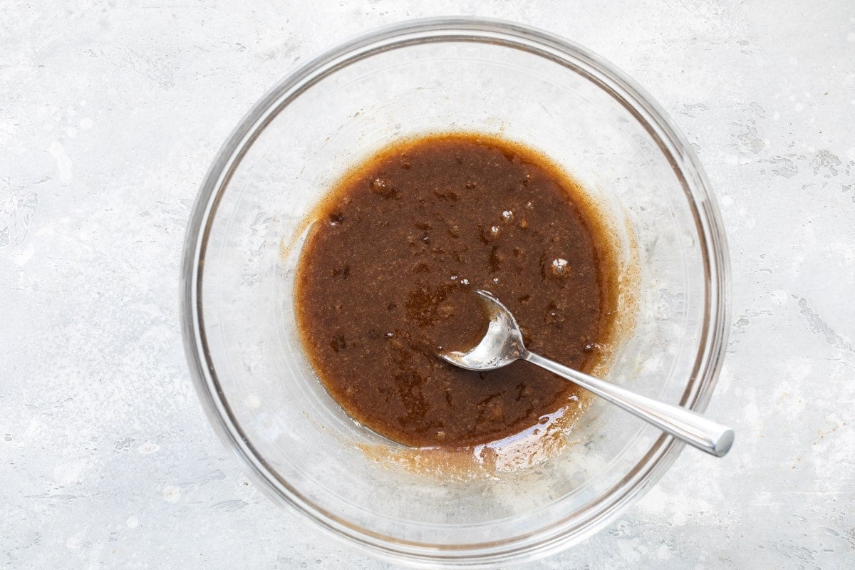 Granola coating liquid in a clear bowl with a spoon resting in it.