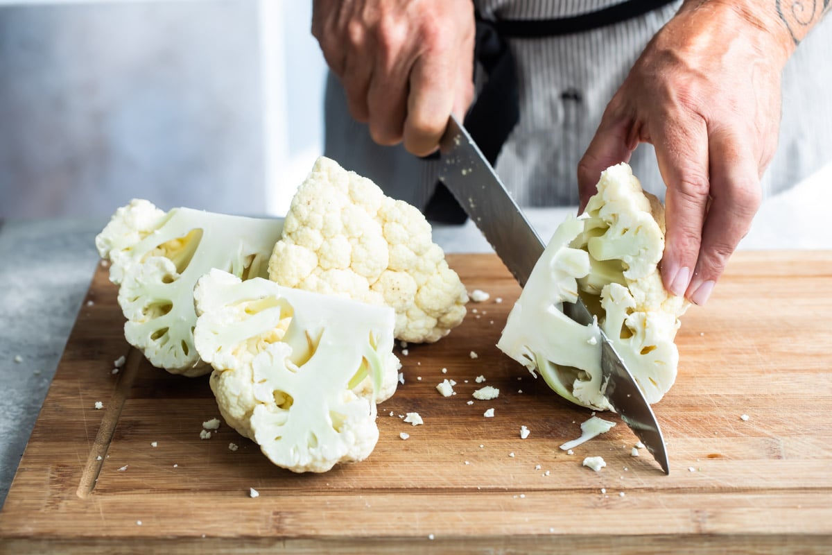 Raw cauliflower being chopped on a wooden cutting board.