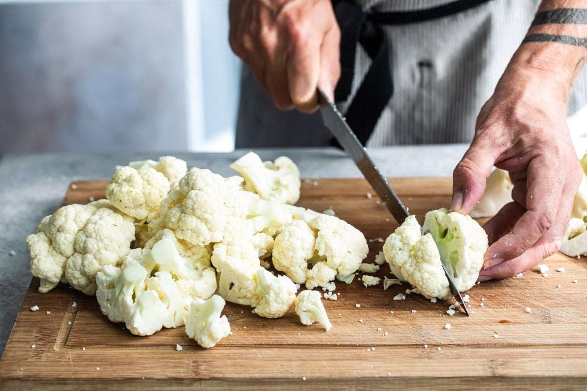 Raw cauliflower being chopped on a wooden cutting board.