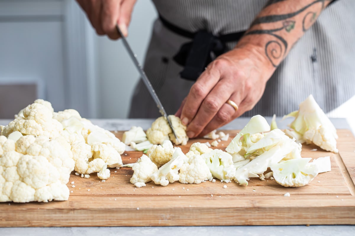 Raw cauliflower being chopped on a wooden cutting board.