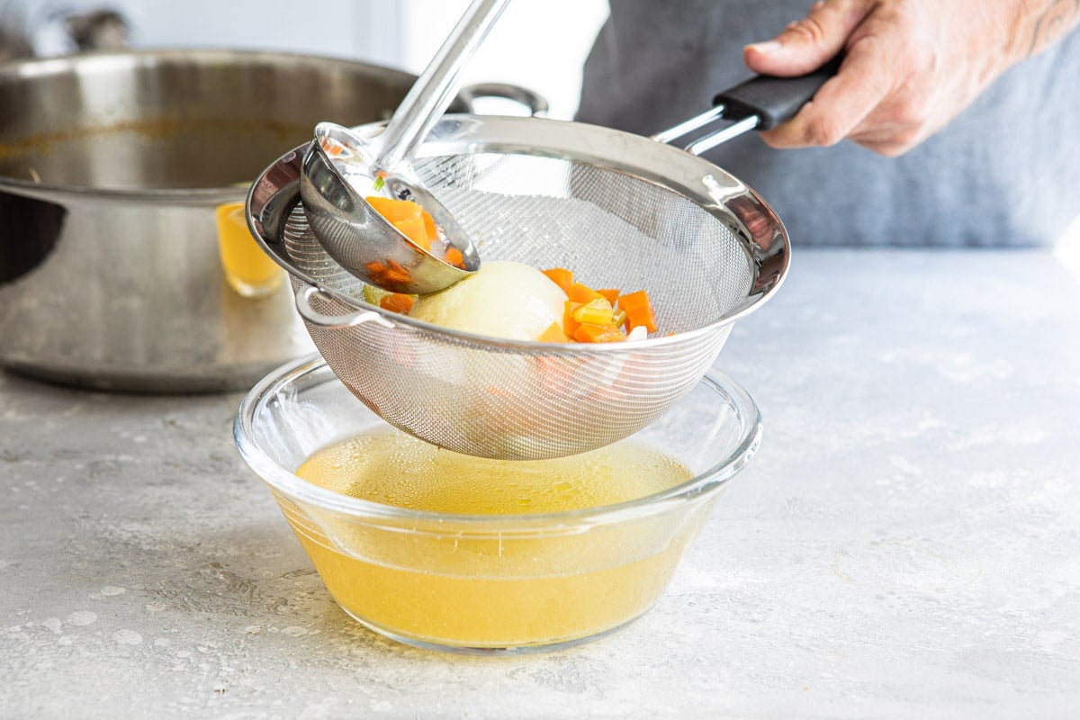 Chicken broth ingredients in a strainer over a clear bowl of chicken broth.