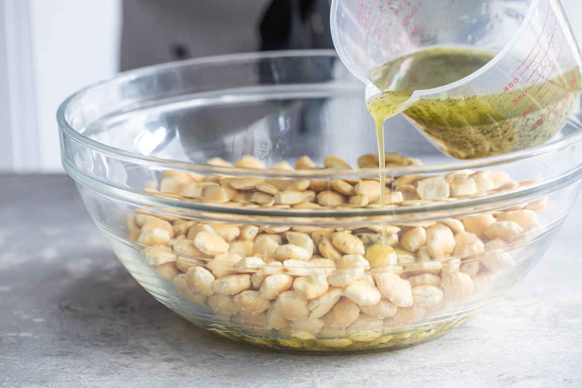 Ranch liquid mixture being poured over oyster crackers in a clear bowl.