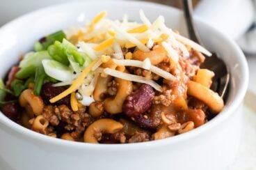 Chili mac topped with cheese, scallions and sour cream in a white bowl.