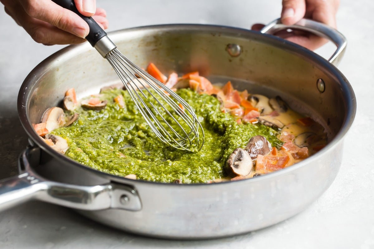 Stirring pesto into a skillet with mushrooms and tomatoes.