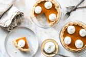 A slice of mini pumpkin pie on a white plate with two mini pumpkin pie and whipped cream behind it.