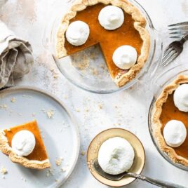 A slice of mini pumpkin pie on a white plate with two mini pumpkin pie and whipped cream behind it.