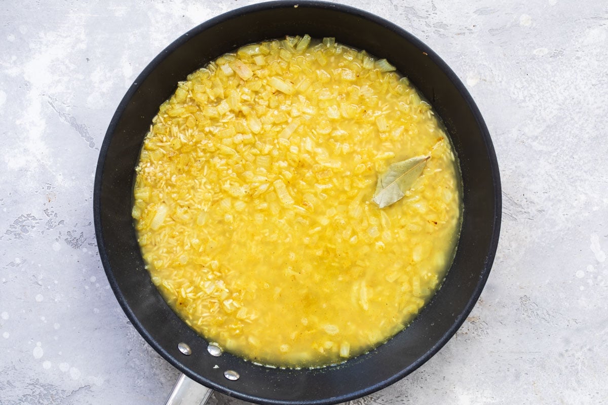 Yellow liquid, rice, onions and a bay leaf in a black skillet.