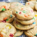 A dozen M&M cookies on a wire rack with a glass of milk and a bowl of M&Ms in the background.