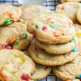 A dozen M&M cookies on a wire rack with a glass of milk and a bowl of M&Ms in the background.