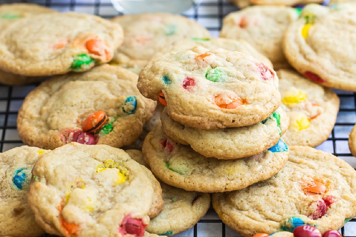 A dozen M&M cookies on a wire rack with a glass of milk and a bowl of M&Ms in the background.