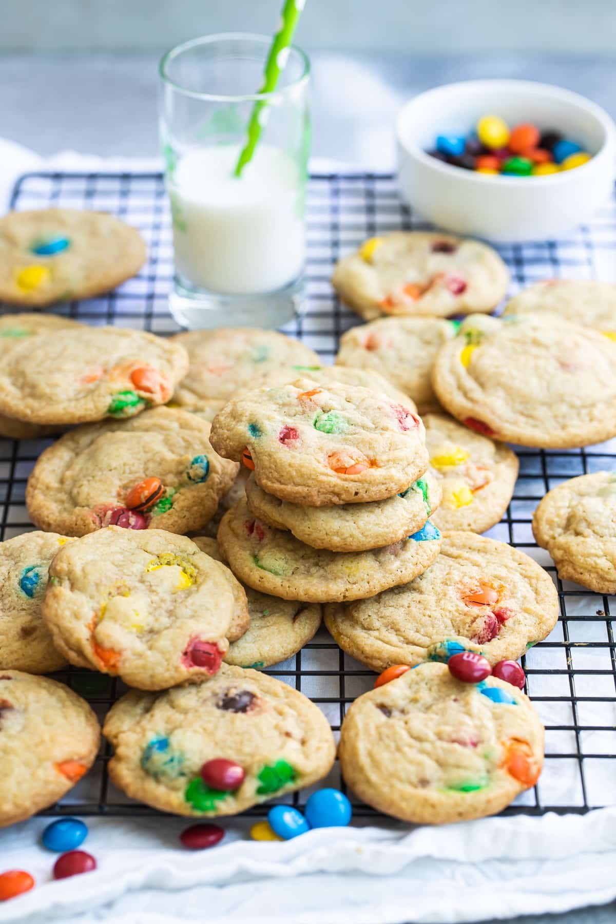 A dozen M&M cookies on a wire rack with a glass of milk and a bowl of M&Ms in the background.