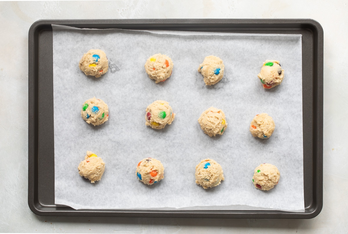 M&M cookie dough balls on a parchment paper lined baking sheet.