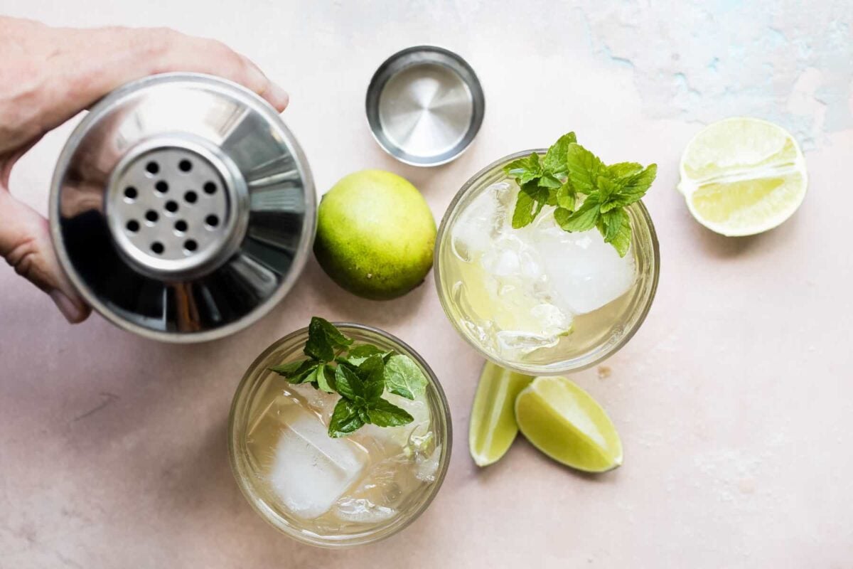 Two Moscow Mules in glasses surrounded by limes and a cocktail shaker.