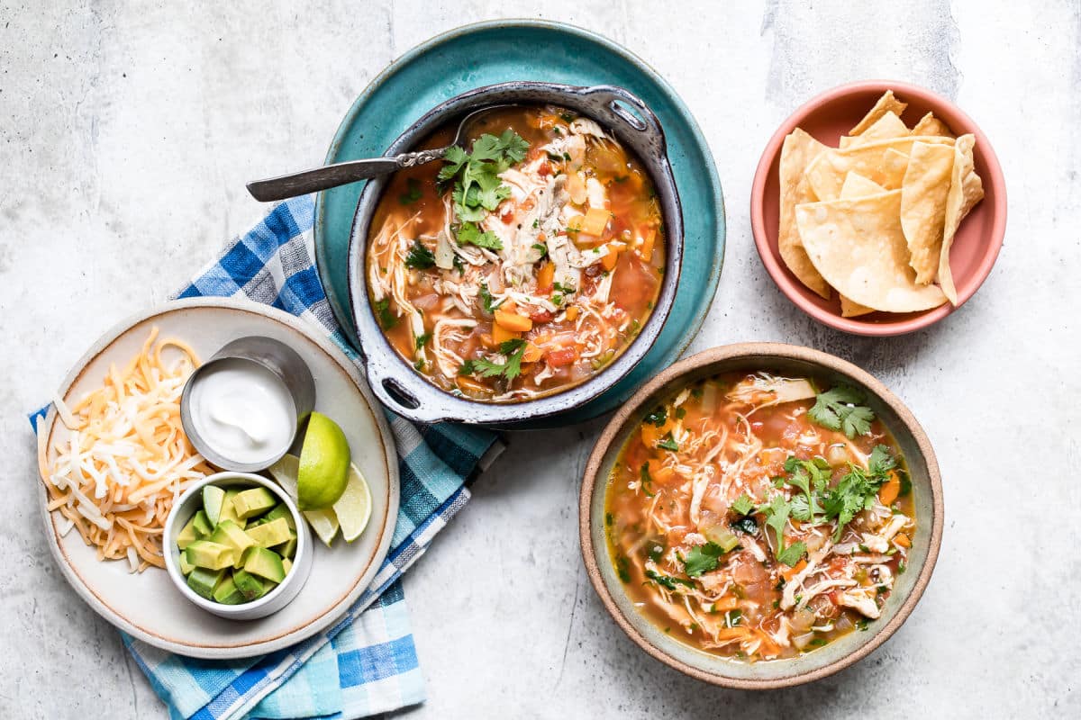 Two bowls of Mexican chicken soup with toppings nearby.