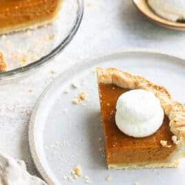 A slice of mini pumpkin pie on a white plate with a mini pumpkin pie and whipped cream behind it.