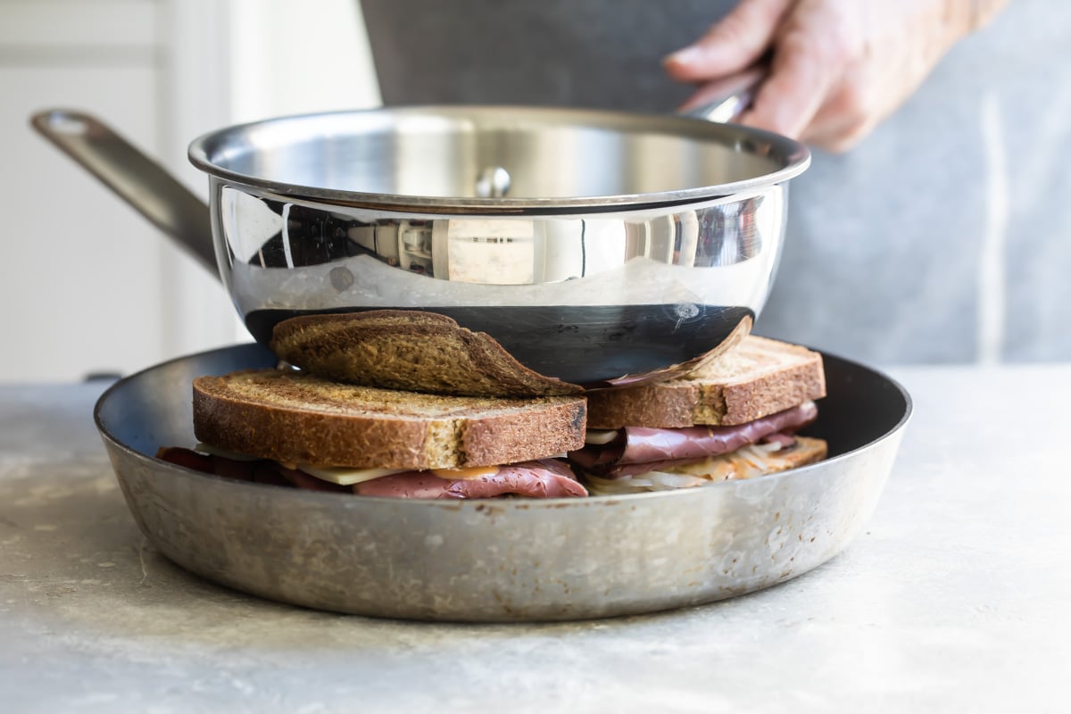 Two reuben sandwiches in a frying pan with a second frying pan pressing on the top of the sandwiches.