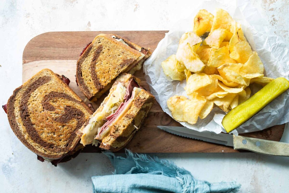 A reuben sandwich on a cutting board surrounded by chips and a pickle spear.