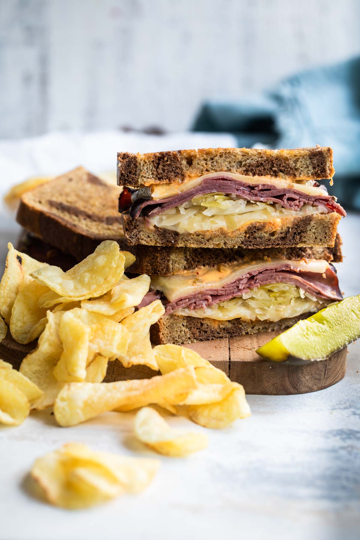A A reuben sandwich on a cutting board surrounded by chips and a pickle spear.